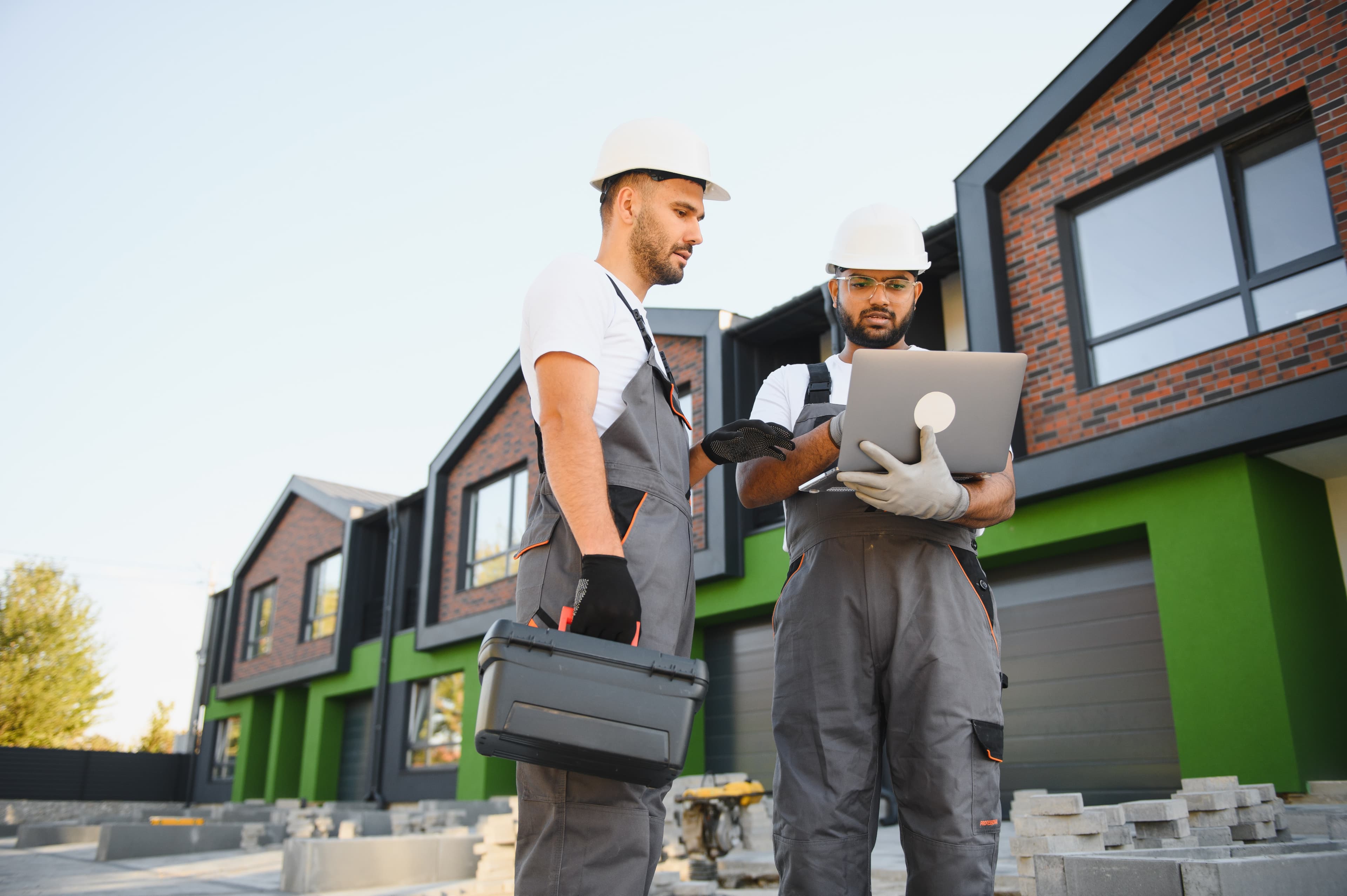 Two builders in hard hats and overalls, one carrying a toolbox, check a laptop outside a newly built row of homes with paving stacked on site