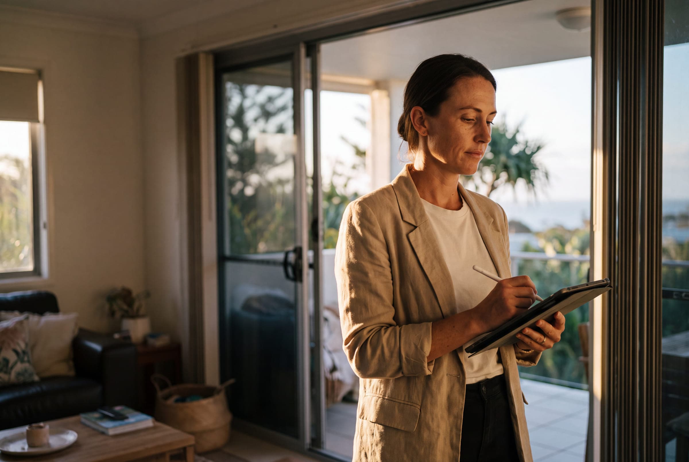 A woman in a linen blazer checking figures on a tablet in the living room of a rental home, with the balcony door open behind her